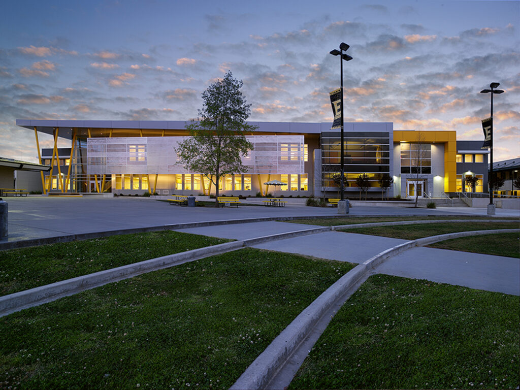 A modern school building with large windows is illuminated at dusk. The sky shows scattered clouds, and banners hang from light poles. Sidewalks and grass are visible in the foreground.