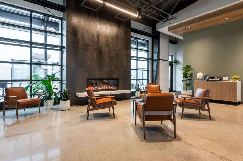 Modern lobby with brown leather chairs, a glass coffee table, electric fireplace, potted plants, large windows, and a refreshment counter on polished concrete flooring. Bright, inviting, and contemporary interior design.