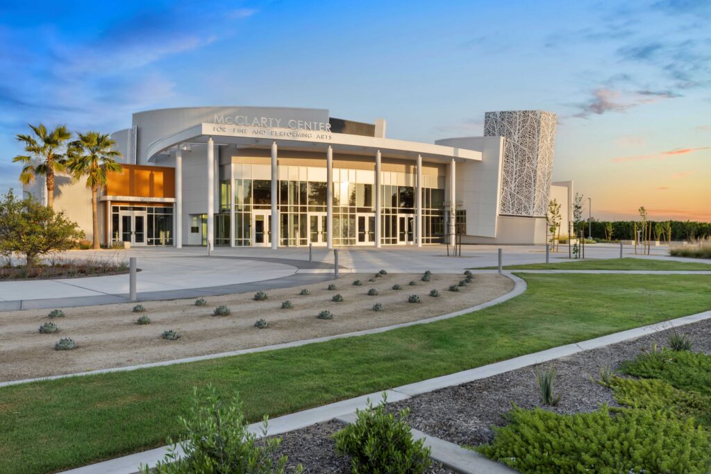 Modern building labeled McClarty Center for the Fine and Performing Arts with large glass windows, palm trees, landscaped paths, and a blue sunset sky in the background.