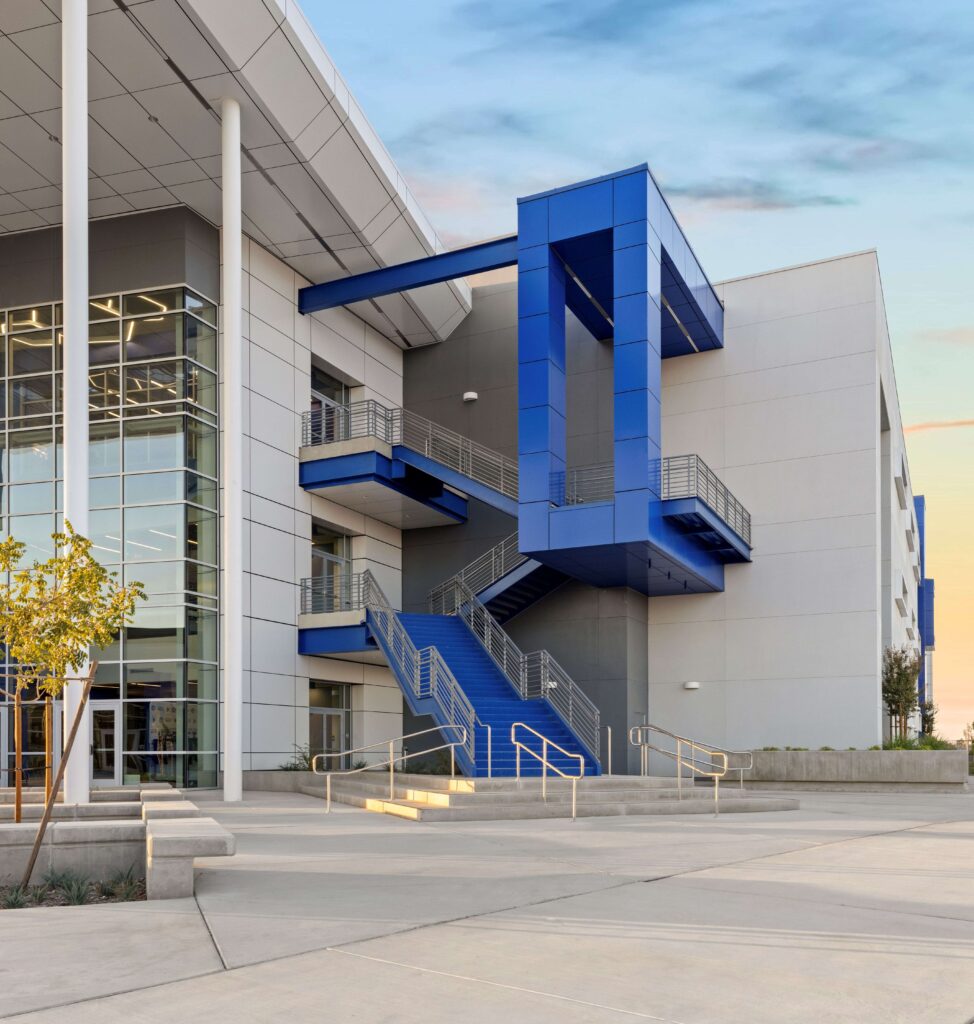 A modern building with blue exterior staircases, large glass windows, and a covered entrance, seen at sunset with a clear sky and minimal landscaping.