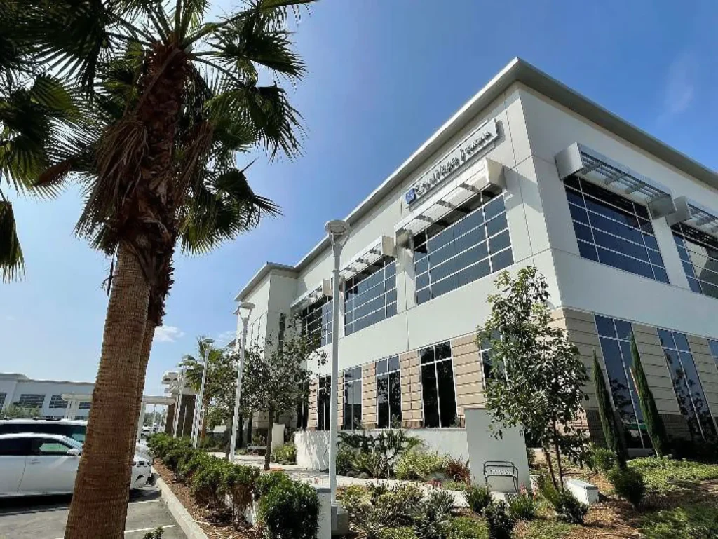 Modern office building with reflective windows, palm trees, and landscaped greenery under a clear blue sky; cars are parked nearby.