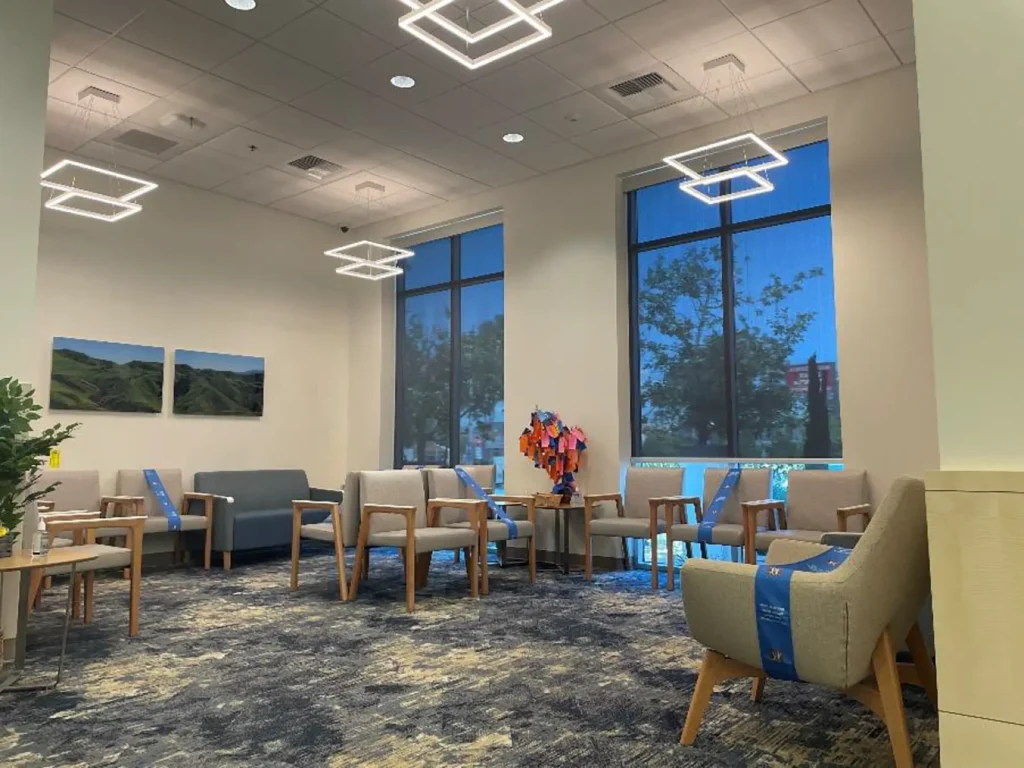 A modern waiting room with beige chairs, blue accents, large windows, geometric ceiling lights, and nature artwork on the wall. Some chairs have blue tape marking social distancing.