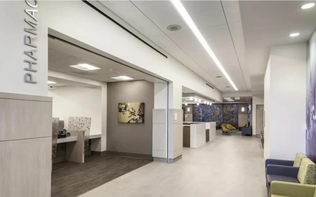 Modern pharmacy interior with light walls, a sign reading PHARMACY, seating area with chairs, artwork on the walls, and bright overhead lighting. The space is clean and organized.