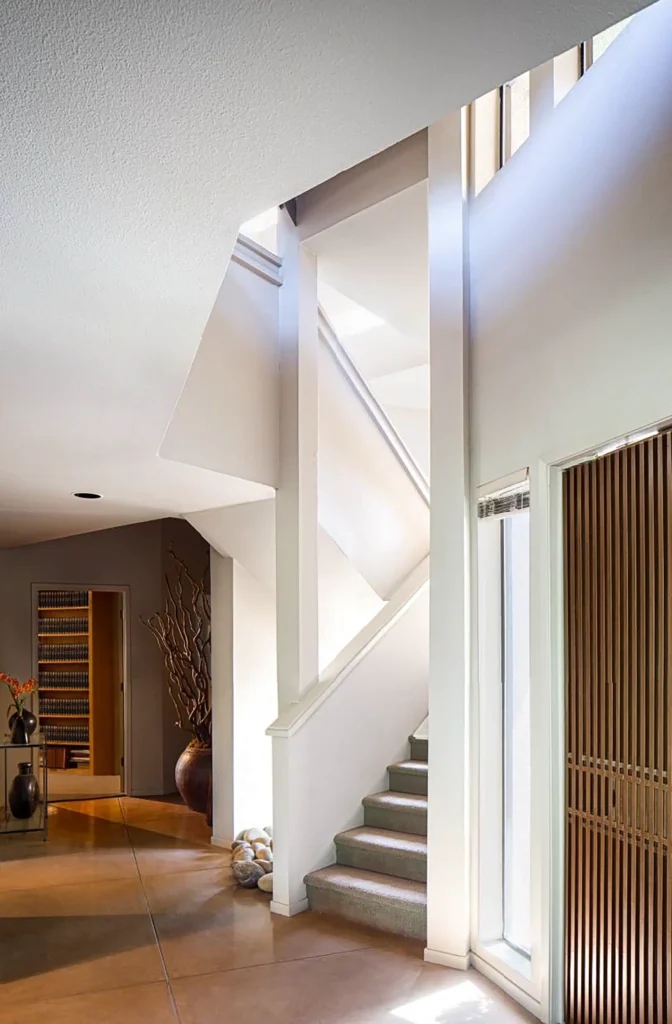 Modern interior with sunlight streaming onto a staircase. Light walls, wooden accents, and plants enhance the clean, airy atmosphere. Stairs lead upward beside large vertical windows.