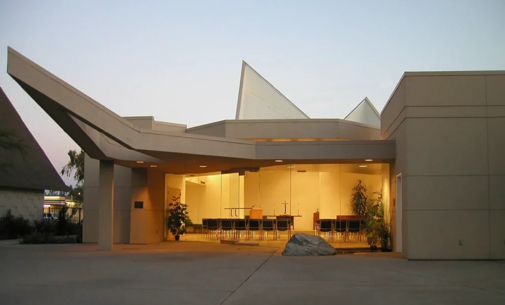 Modern building with angular rooflines, large windows, and warm interior lighting. Several chairs are arranged inside, facing a podium, with plants and a large rock outside near the entrance at dusk.