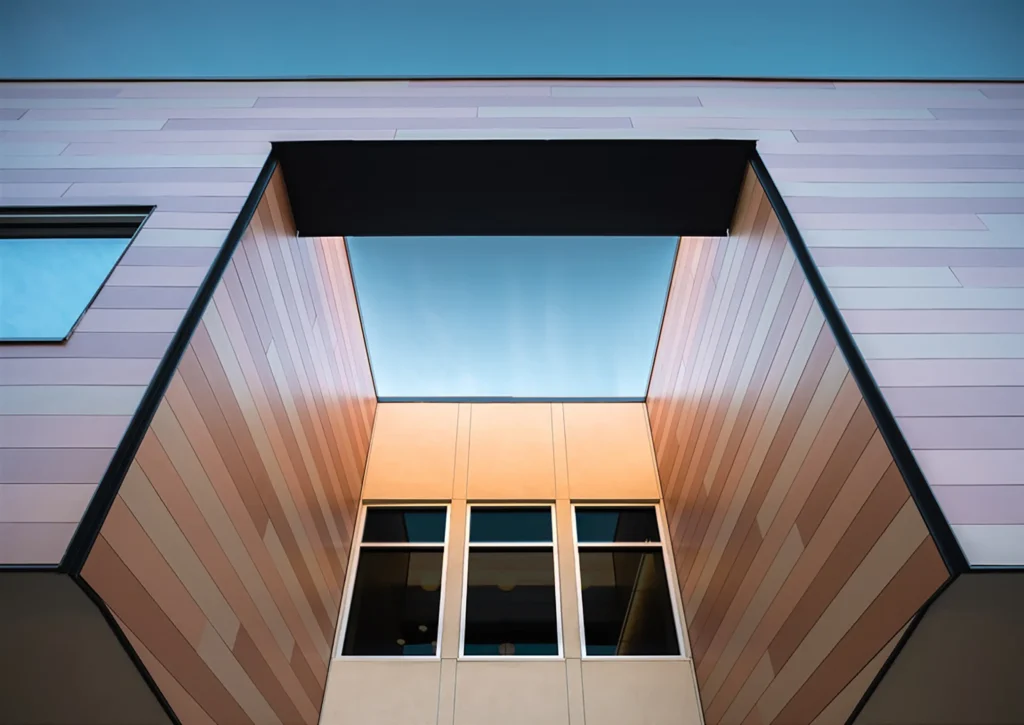 Modern building facade with large windows and pinkish paneling, viewed from below. The structure frames a rectangular opening revealing blue sky.