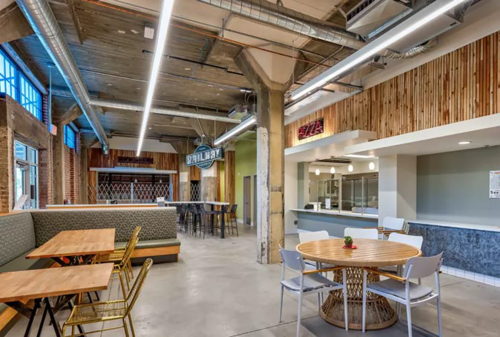Modern indoor dining area with wooden tables, mixed seating, exposed concrete and pipes, and food counter signs for Pizza and Grill Bar. Large windows provide natural light.