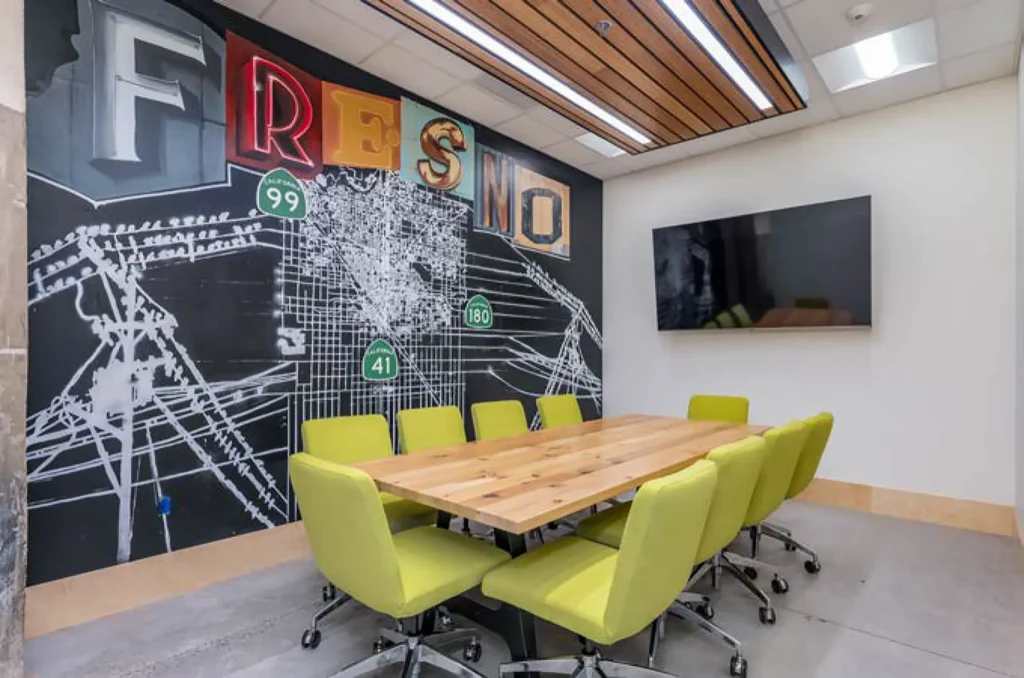 A modern conference room with a wooden table, eight green chairs, a wall mural reading Fresno, a city map, highway signs, and a large flat-screen TV.