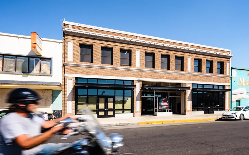 A motorcyclist rides past a two-story brick building with large windows on a sunny day.