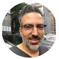 A man with gray hair, glasses, and a beard smiles outdoors in a city setting, with cars and tall buildings in the background.
