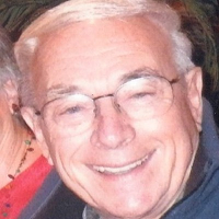 Older man with short white hair and glasses smiling warmly at the camera, wearing a dark jacket. The background is partially obscured and another person is barely visible to his left.
